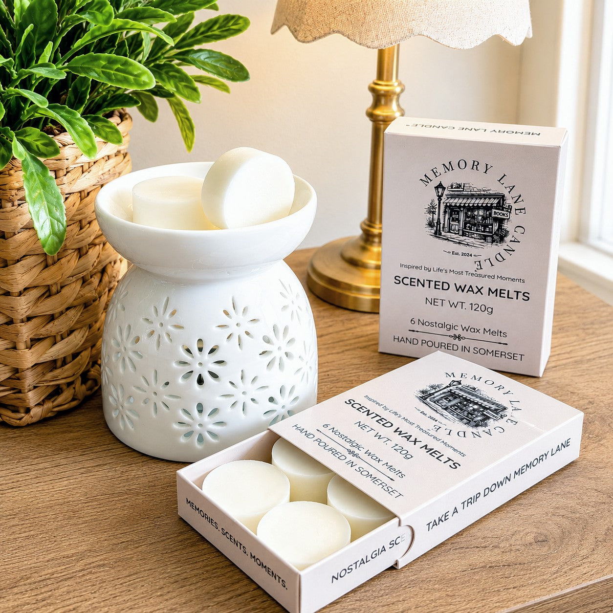 A boxed set of Memory Lane Candle scented wax melts displayed on a wooden table beside a white ceramic wax warmer and a potted plant. The open box shows round off-white wax melts, and the branded retail box and another closed box stand behind them.