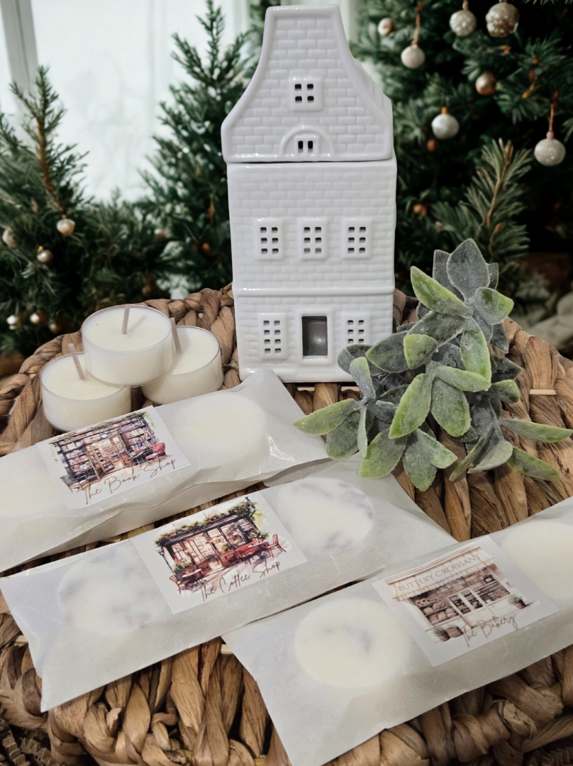 Ceramic wax burner with melts & candles and decorative elements on a woven surface with Christmas trees in the background.