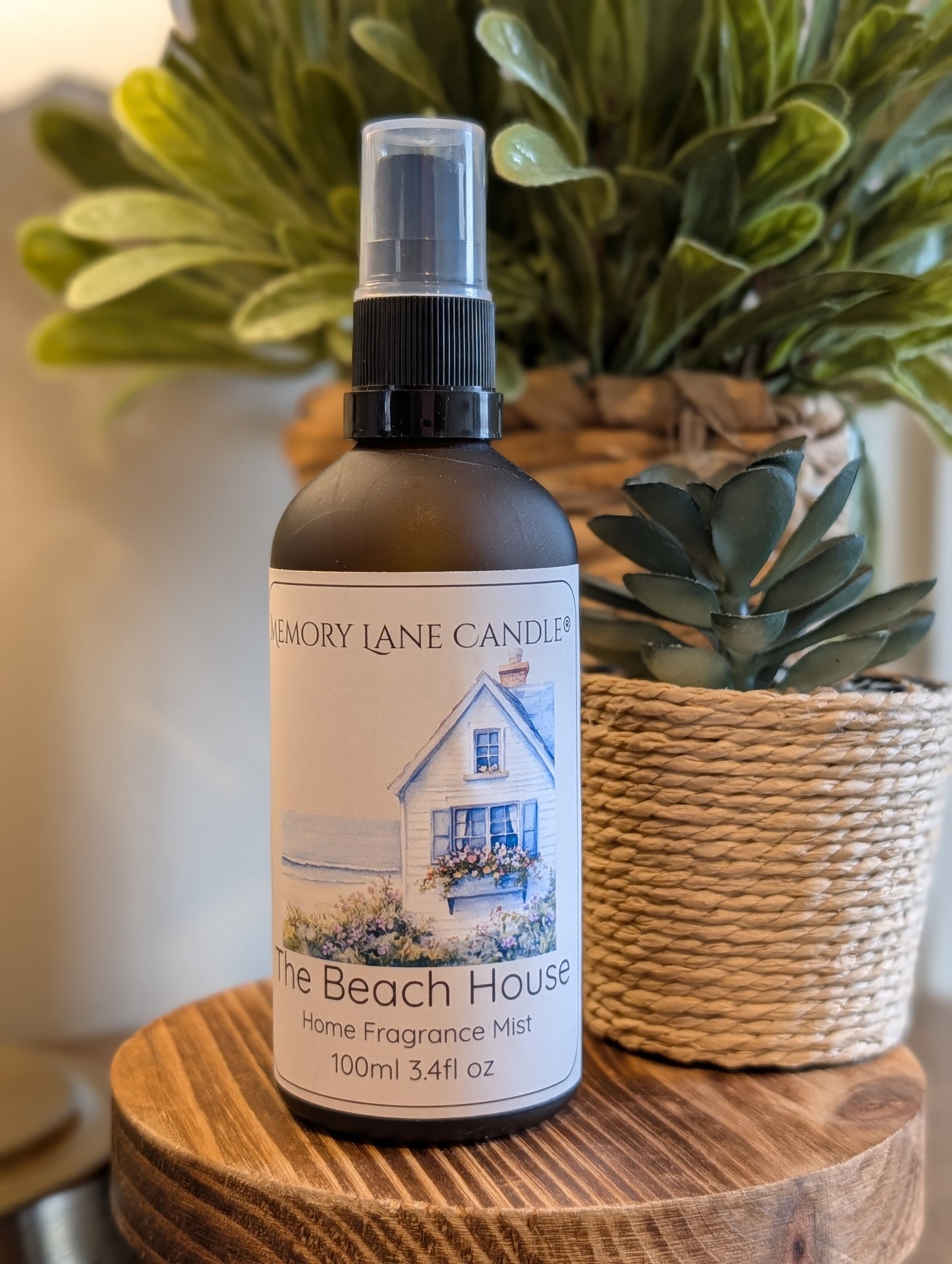 Bottle of 'The Beach House' home fragrance mist by Memory Lane Candles on a wooden surface with plants in the background.