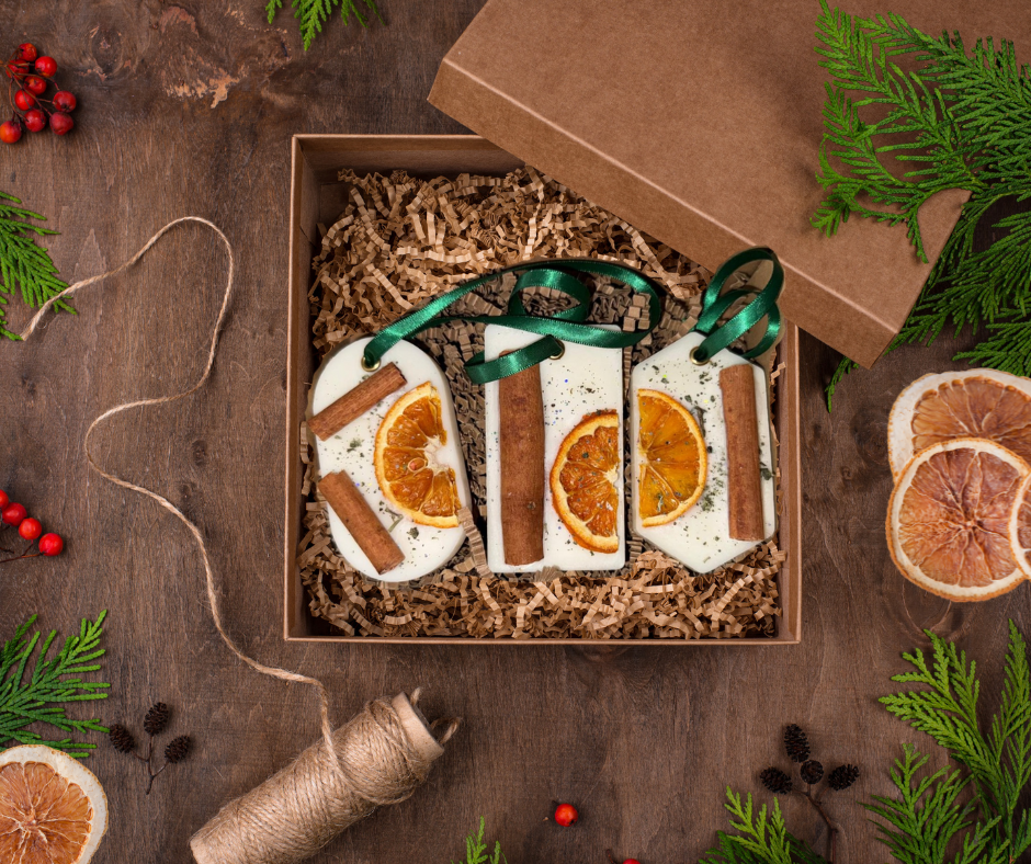 A set of three festive wax melt hangers displayed in a kraft gift box, each decorated with dried orange slices, cinnamon sticks, and green ribbon loops. The box is filled with shredded kraft paper and surrounded by Christmas greenery, pinecones, red berries, and candy canes for a cosy, rustic holiday feel.