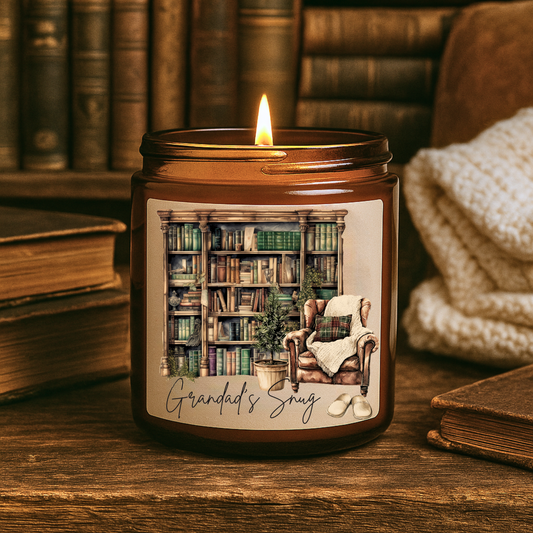 Candle with a design of a cozy room and 'Grandad's Snug' text, placed on a wooden surface with books and a blanket in the background.