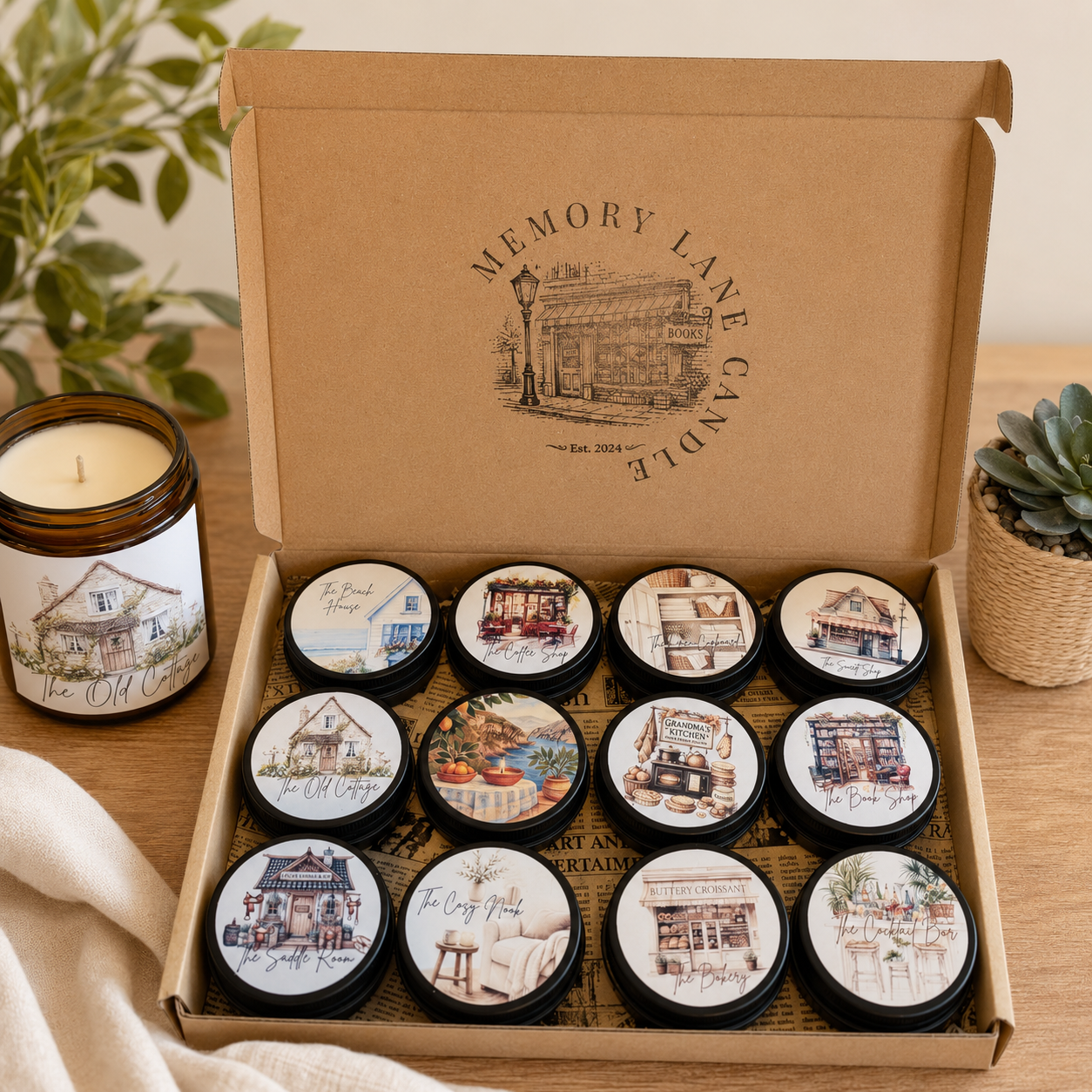 An open kraft cardboard gift box labelled 'Memory Lane Candle' holds 12 small round tin candles with illustrated labels. A larger amber glass jar candle sits to the left on a wooden table, with a cream cloth and potted greenery in the background, creating a warm home-style product presentation.