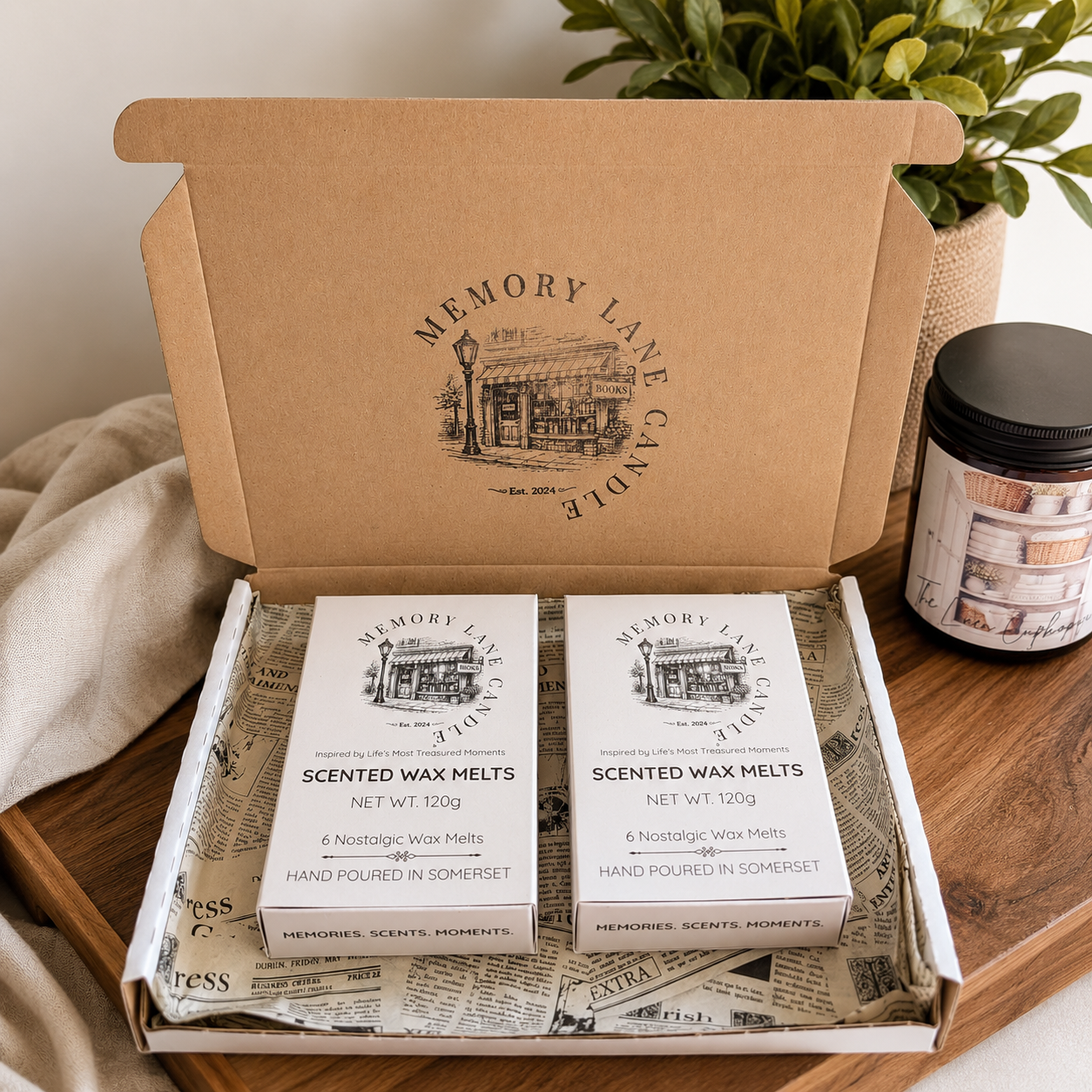 An open kraft cardboard gift box from Memory Lane Candle sits on a wooden surface, displaying two boxed scented wax melts wrapped in white paper with black printed illustrations. A matching candle jar and a beige textile are visible in the styled setting.