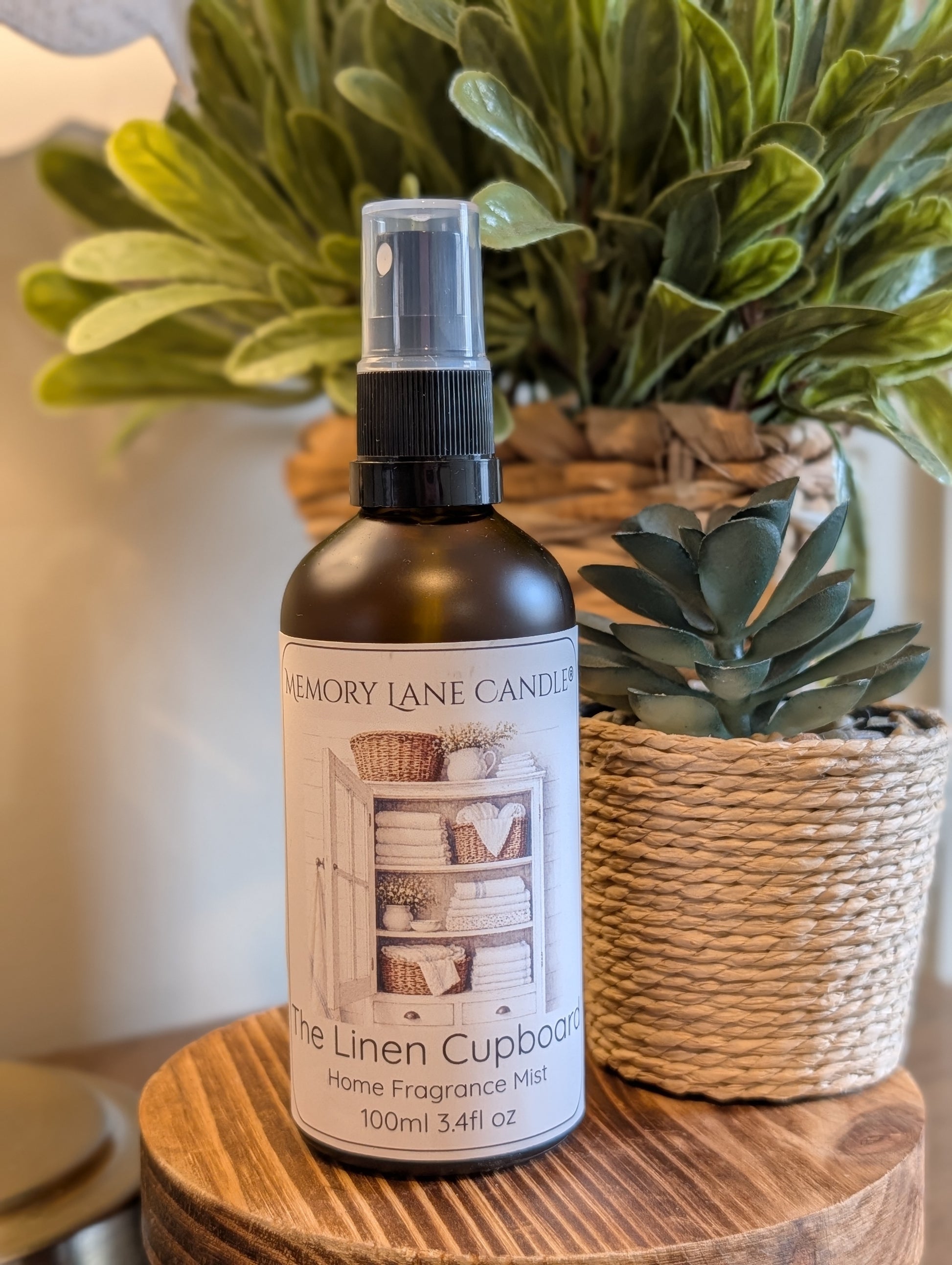 Bottle of Memory Lane Candle Co. linen cupboard fragrance mist on a wooden surface with plants in the background.