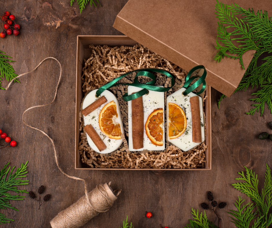A set of three festive wax melt hangers displayed in a kraft gift box, each decorated with dried orange slices, cinnamon sticks, and green ribbon loops. The box is filled with shredded kraft paper and surrounded by Christmas greenery, pinecones, red berries, and candy canes for a cosy, rustic holiday feel.