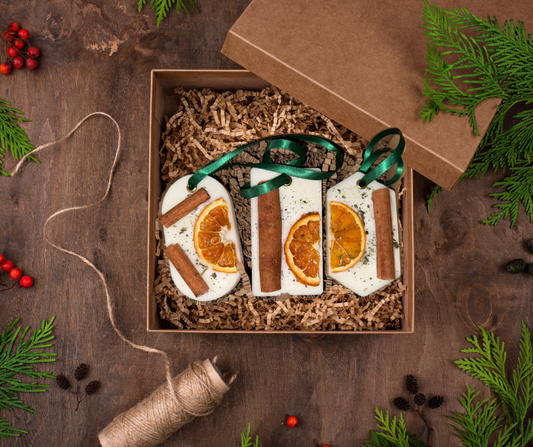 A set of three festive wax melt hangers displayed in a kraft gift box, each decorated with dried orange slices, cinnamon sticks, and green ribbon loops. The box is filled with shredded kraft paper and surrounded by Christmas greenery, pinecones, red berries, and candy canes for a cosy, rustic holiday feel.